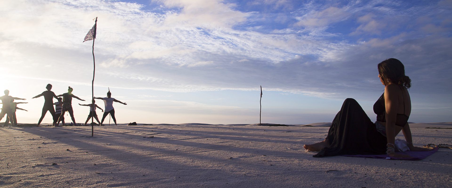 A girl looking at a group of people doing yoga on the sand dunes in Atins